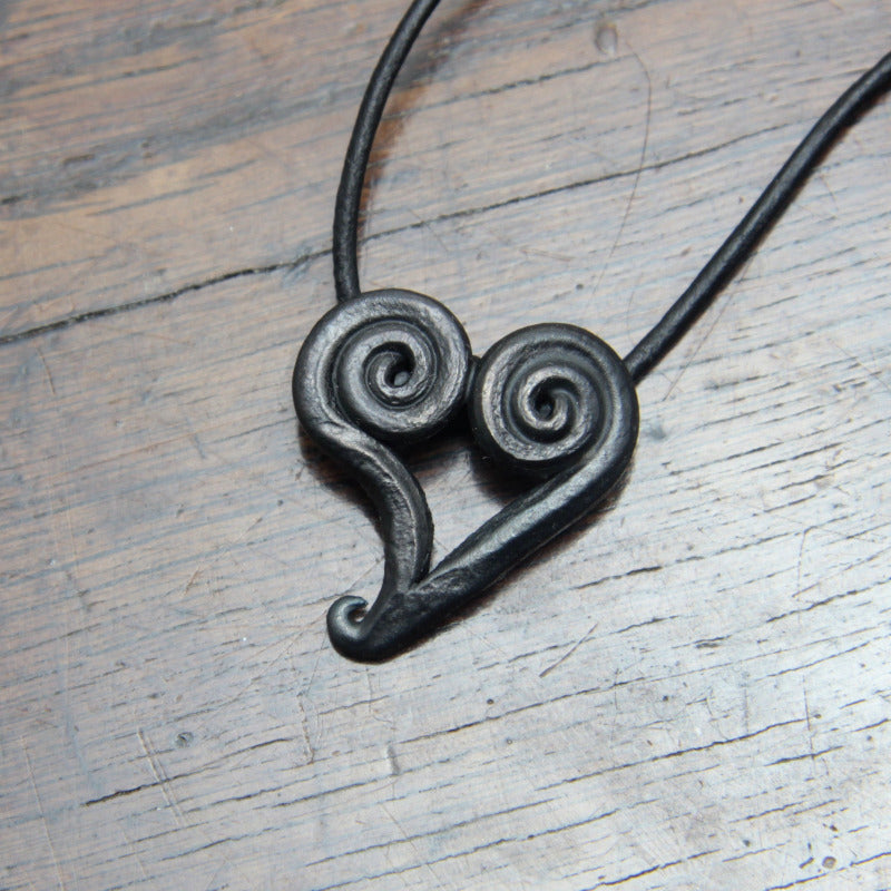 Hand Forged Black Iron Spiral heart pendant, made and designed by M Barran at Taitaya Forge. On a black leather cord, photographed on a dark wood surface. 
